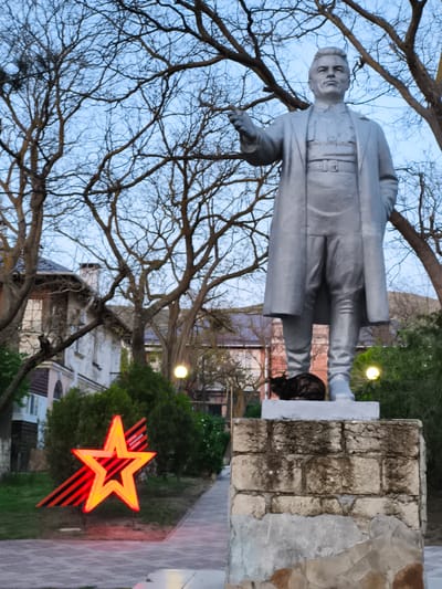 A statue of a man in a long coat stands in a park in Орджоникидзевский поселковый совет, Ukraine. The statue is made of stone or metal and stands on a brick pedestal. A dark-colored cat is sitting on the pedestal at the base of the statue. In the background, there is a building with several stories and bare trees. To the left of the statue, there is a red star shape with black and orange stripes behind it.