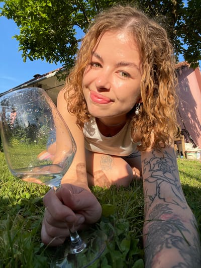 The image captures a young woman lying on green grass, looking directly into the camera with a gentle smile. She has curly, light brown hair, fair skin that appears dewy in the sunlight, and light-colored eyes. She wears a light-colored tank top, a pearl earring, and a thin necklace. Her arms and legs are adorned with several tattoos, including a large, intricate design resembling wings or an abstract pattern on her upper arm and a rose on her lower leg.

In the foreground, she holds a clear, stemmed glass, likely a wine glass, filled with a transparent liquid and showing droplets on its exterior. The glass is positioned close to the camera, slightly obscuring part of her body and the background.

The setting is outdoors on a bright, sunny day. She is surrounded by vibrant green grass. In the background, a light pink building wall is visible, with a clear blue sky and lush green tree foliage above. The sunlight suggests it is daytime, creating a warm and serene atmosphere.

A small, faint piece of Cyrillic text, "ПИВО" (PIVO, meaning 'beer'), is visible on a distant wooden object or crate in the background. The scene is located in Danilovgrad, Montenegro.
