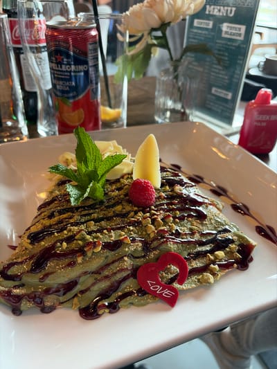 The image shows a dessert crepe, possibly a pistachio crepe, on a white plate. It is topped with a dark sauce, possibly chocolate, chopped pistachios, a raspberry, a slice of banana, white chocolate, and a sprig of mint. A red heart-shaped decoration with the word "LOVE" on it rests on the edge of the crepe. The background includes cans of Pellegrino Aranciata Rossa and possibly other beverages, along with a menu from "Geek Bar." The location is likely in Willowbrook, United States of America, based on the context of the menu and the presence of the American brand Geek Bar.