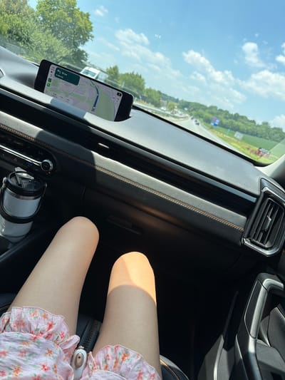 This is a photograph taken from inside a vehicle, looking forward and slightly to the right. The perspective shows the dashboard, a smartphone mounted on the dashboard displaying a navigation app, a cup holder with a white tumbler, and a portion of the passenger's legs and dress. The exterior view through the windshield shows a highway with other vehicles, trees, and a bright blue sky with scattered clouds. Based on the navigation displayed on the phone, the location is East Hempfield Township, United States of America. The time shown on the phone is 4:38 PM. The speed limit indicated on the navigation is 55 mph.