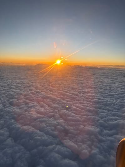 The image shows a sunrise or sunset viewed from above the clouds. The sun is positioned on the left, emitting bright rays, and the sky transitions from blue to orange near the horizon. A vast expanse of clouds, textured with individual billows and shadowing, stretches out to the right side of the frame. The scene does not depict any specific identifiable location. There is no explicit indication of the image being taken in the city of Vecsés, Hungary. The image appears to have been rotated 90 degrees counterclockwise.