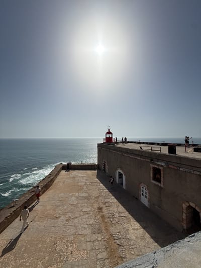 The image captures a sunlit scene at the Fortaleza de Nazaré in Nazaré, Portugal, featuring a weathered stone courtyard on the cliffside. A lighthouse with a red top stands prominently on the building's roof. Several people are visible in the area. The Atlantic Ocean extends to the horizon, with visible waves crashing against the cliffs. The sky is bright with the sun high.