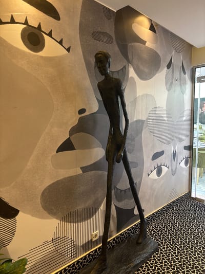 A dark, slender human sculpture stands against a wall decorated with a large, abstract mural featuring stylized eyes and geometric patterns in shades of gray and beige. The floor is covered with a black and white patterned carpet. To the right, a glass door reveals a dining area with tables and chairs, and a glimpse of patrons. The scene is set in Khurba Souq, Jordan.