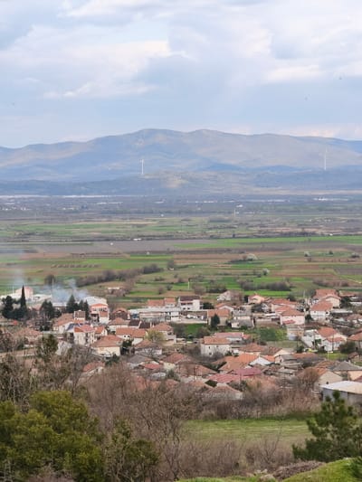 An elevated, wide-angle view captures the village of Negorci, North Macedonia, situated in a valley, with agricultural fields extending into the midground and mountains defining the horizon under a cloudy sky.

The foreground consists of unkempt earth, dry brown brush, and scattered green foliage, including a small group of green trees on the left and a pine branch on the far right.

The midground is dominated by the village of Negorci, characterized by numerous residential buildings with predominantly red or terracotta tiled roofs and light-colored facades. Several plumes of light grey smoke are visible rising from multiple points within the village, suggesting active chimneys or outdoor burning. Beyond the village, a vast expanse of agricultural land is visible, segmented into rectangular plots displaying varying shades of brown (tilled earth) and green (early crops or grass). Sparse trees and shrubs are scattered throughout these fields.

The background features a range of hazy, blue-grey mountains stretching across the entire horizon. Two prominent white wind turbines are observed on the distant mountain ridges, one to the left of center and another to the right of center. The sky above is partially covered by white and grey clouds against a light blue backdrop. No individuals are visible in the scene.