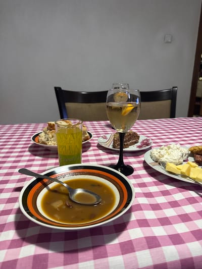 An indoor scene in Erma Reka, Bulgaria, depicts a dining table set for a meal. The table is covered with a pink and white checkered tablecloth. In the immediate foreground, a white bowl with orange and black concentric stripes contains a brown soup or stew with visible solid components; a silver spoon rests within the bowl. To the left, a clear, tall glass with "APPLE" text printed on its side holds a green liquid. Behind it, a small white plate contains pieces of white bread. To the right, a stemmed wine glass with a black stem holds a light-colored liquid with a lemon slice, with a second, blurry similar glass visible behind it. Adjacent to this, a small white plate with a floral pattern presents a slice of chocolate layer cake. Further to the right, a larger white platter contains a portion of white creamy salad, cubed yellow cheese, and slices of red processed meat (salami or sausage); a silver fork is positioned on this plate. The background features a plain light gray wall with a single white light switch or outlet. Part of a dark wooden dining chair with a beige upholstered back is visible behind the table. A dark, blurry vertical object, possibly furniture, is on the far right edge of the frame. No individuals are present.
