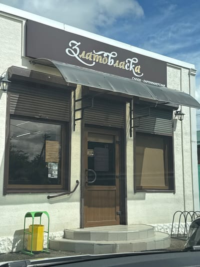 The image shows a three-story building in Yelizavetinskaya, Russia. The sign on the awning reads "Златовласка", with "САЛОН - ПАРИКМАХЕРСКАЯ" (salon - hairdresser) written below. The building has brown framed windows. The two lower windows have what appear to be security shutters partially closed. The second story window has a small sign inside. A metal handrail is located below the second-story window and a green and yellow trash bin is located to the right of the bottom window.