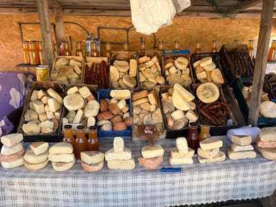 At a market stall in Ponikvica, Montenegro, a variety of cheeses and bottled beverages are displayed for sale. The stall features wooden shelves and panels, with a checkered tablecloth covering the display surface. Large crates overflow with numerous pieces of cheese of various shapes, sizes, and textures, some appearing aged and crumbly, others smooth and firm. Several boxes are filled with dark, cured sausages. On the shelves above, rows of clear glass bottles containing amber-colored liquids, likely juices or preserves, are arranged. Water bottles and more juice bottles are also visible. In the foreground, stacks of cheese are arranged on the tablecloth, with a blue-handled knife resting on the fabric. The overall scene suggests a local market offering artisanal food products.