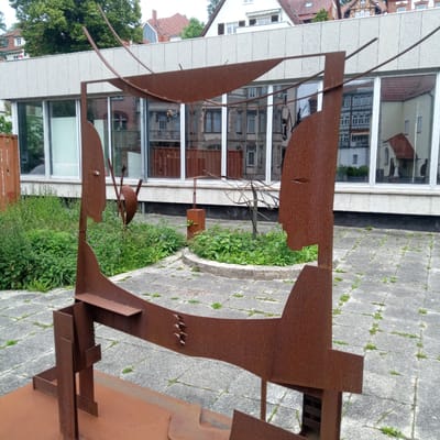 An abstract, rusty metal sculpture is situated in an outdoor courtyard with a modern building featuring large glass windows in the background. The sculpture is composed of various flat and curved metal pieces, forming a cage-like structure with abstract, face-like elements. The courtyard pavement is made of grey square tiles interspersed with patches of green vegetation. The building in the background has a grey speckled facade above the windows, and the reflections in the glass show other buildings with red tiled roofs and windows. Lush green plants and small wildflowers are growing in a raised circular bed in the middle ground, and further to the left, more greenery is visible. The sculpture appears to be permanently installed, given its scale and material. The scene is likely in a public space, possibly an art installation or part of a museum's exterior. The overall impression is one of industrial art meeting natural elements and urban architecture. The image was taken in Eisenach, Germany.