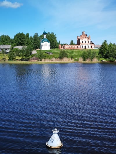A vibrant outdoor scene captures a wide body of dark blue, rippled water in the foreground, likely a lake or river. In the lower right, a white, conical navigational buoy floats, marked with the dark blue number "19" and featuring a crown-like top structure.
The far bank is lush and green, covered with grass, reeds, and dense deciduous trees. Two prominent architectural structures stand on a rise along this bank. To the left is a small, well-maintained white chapel with a striking green, onion-shaped dome topped with a cross, characteristic of Russian Orthodox architecture. Behind and to the left of the chapel, several rustic wooden houses with grey roofs are partially visible amidst the foliage.
To the right of the chapel, further along the bank, are the substantial ruins of a large red brick building, possibly an older church or monastery. This structure shows significant signs of decay, with crumbling walls, exposed brickwork, and large sections of its roof missing, though remnants of arched windows and a dome base are still discernible.
The sky above is a clear, bright blue with a few wispy white clouds, indicating a sunny day. The scene evokes a sense of history and tranquility, juxtaposing a preserved sacred building with an ancient ruin, all within a natural setting. The time of day is bright daylight. The location is Анхимово, Russia.