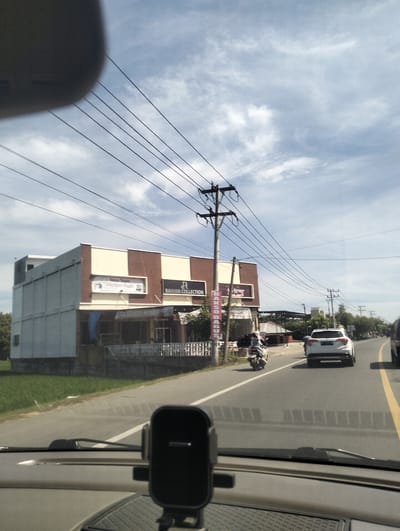 The image captures a street scene in Teungoh Baro, Indonesia, on a bright, cloudy day. A white car is driving down the road, followed by a motorcycle. The road is paved and marked with yellow and white lines. To the left of the road, there is a building with a sign that reads "RAIHAN COLLECTION". There are also several utility poles with wires running across the scene. The image appears to be taken from inside a vehicle, with a car phone mount visible in the foreground.