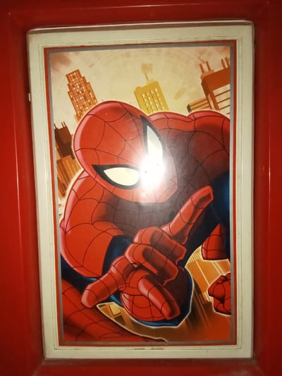 This image captures a framed illustration of the Marvel Comics character Spider-Man, situated in Ciputri, Indonesia. The illustration prominently features Spider-Man's upper body and arms, rendered in his classic red and blue costume with black web detailing and distinct white eye lenses. His right hand is raised, with the index and middle fingers extended in a characteristic web-shooting posture, while his left hand is also visible and similarly positioned. The background behind the character is a stylized urban landscape, depicting brown and yellow high-rise buildings against a light-toned, possibly sky-like, setting with subtle circular patterns. The illustration is encased within a white frame that has a thin silver-grey inner border. This framed piece is set into a larger, uniformly red, textured surface, possibly part of a panel or enclosure. A notable glare is present near the center-right of Spider-Man's mask.