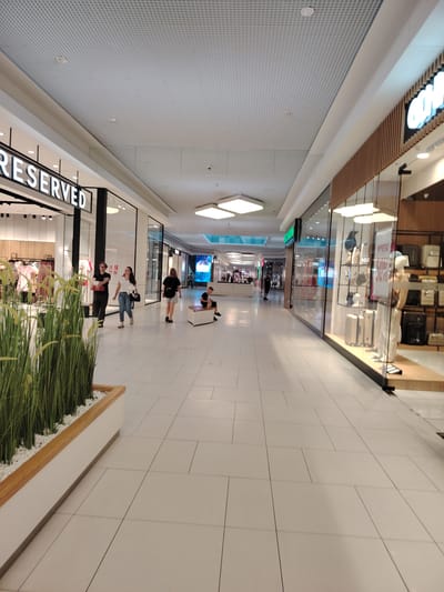 The image depicts an indoor shopping mall with a modern, well-lit interior. The scene is captured from a low-angle perspective, looking down a wide, tiled walkway. On the left, a store with the prominent "RESERVED" signage is visible, showcasing clothing racks inside. In the foreground, a planter filled with tall green grass adds a touch of nature to the sterile environment.

Several people are present, walking along the corridor. Two individuals, a man and a woman, are walking together towards the viewer, carrying bags. Further down the mall, another woman in a black t-shirt and shorts is walking away. In the distance, a young boy sits on a bench, looking at his phone.

The mall's architecture features a grid-patterned ceiling with recessed lighting and square, illuminated ceiling panels. The floor is made of large, light-colored tiles with thin dark grout lines. The right side of the walkway is lined with shopfronts, one of which displays mannequins and luggage, with a sign indicating a "WYPRZEDAŻ" (sale) with "50%" highlighted. The overall atmosphere appears calm and everyday, suggesting a typical shopping day. The lighting indicates it is daytime, and the indoor setting means weather is not a factor. The location is identified as Wołomin, Poland.