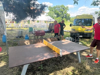 The image depicts an outdoor gathering with people playing ping pong. The scene is set in a grassy area with a ping pong table in the foreground. A yellow van is parked behind the table, and a grill with food is visible further back. There are also tents, tables, and chairs set up, suggesting a picnic or barbecue event.

Three men are visible in the image. One man in a red shirt is holding a ping pong paddle, appearing to be in the middle of a game. Another man in a black t-shirt stands near the grill, while a third man in a red shirt is on the right side of the frame, also holding a paddle.

The weather appears to be sunny with some clouds in the sky, indicating it's daytime. The location seems to be a rural or park-like setting in Lovets, Bulgaria, with trees and vegetation surrounding the area. The overall mood seems relaxed and recreational, with people enjoying the outdoor activity.

A license plate on the yellow van reads "10 CT". Some red crates are also visible near the grill. The ping pong table has a yellow net with white perforated sides. The man in the red shirt on the right is wearing orange athletic shoes and white socks.
