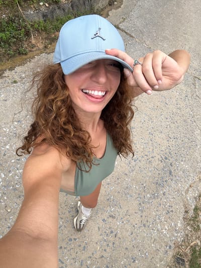 The image shows a woman in Suối Giàng, Vietnam, taking a selfie from a high-angle view. She is wearing a light blue baseball cap with a silver logo, a green top, and one visible running shoe. Her hair is curly and light brown. She is smiling, holding the brim of her cap with her left hand, which has a green ring on one finger. The background is a concrete path or road with some grassy areas visible along the edges.