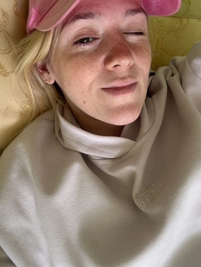 A close-up, overhead shot shows a person with blonde hair and freckles wearing a pink hat and a beige hooded sweatshirt. The person is lying down, propped up by a yellow patterned cushion. They are looking at the camera with one eye open and the other closed, with a slight smile. The sweatshirt has a repeating text logo on the chest that reads "PARIS COUTURE AMOUR L'AMOUR". The background is a yellow patterned fabric, possibly a couch or armchair. The image appears to have been taken indoors, in a well-lit room. There is no indication of the location being in Sarov, Russia.