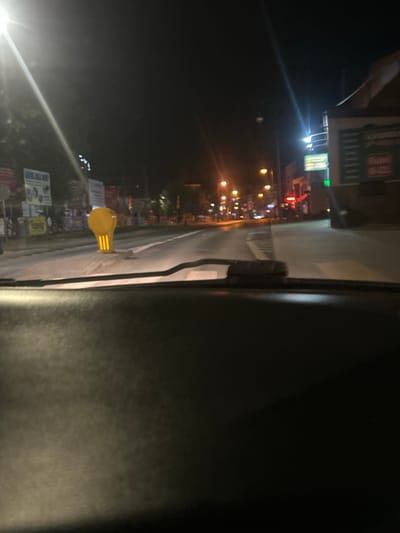 The image is a night-time view from inside a vehicle, looking out at a street in Siedlce, Poland. In the foreground, the dark, textured dashboard of the car is visible. The road ahead is illuminated by streetlights and lights from buildings. A prominent yellow traffic cone with reflective yellow stripes is positioned in the middle of the road. On the left side of the street, there are various signs mounted on poles. On the right side, a building with illuminated signage is present. The overall scene depicts a typical urban street at night, with the traffic cone suggesting ongoing roadwork or a temporary obstruction.