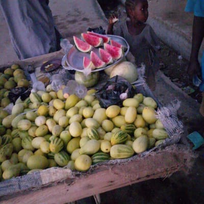 In Wuro Allahamdu, Nigeria, a market scene unfolds with a focus on a stall laden with fruits. A young child, likely female due to visible earrings, sits to the right, wearing a grey knitted garment and looking to the left of the frame. An adult figure, partially obscured by fabric, stands to the left.
The foreground is dominated by a wooden stall covered with a rough, textured fabric. This stall displays a vast quantity of small, oval-shaped fruits, primarily yellow with some green striped varieties, resembling melons or cucumbers. A metal tray on the stall holds several sliced watermelons, their red flesh and white rinds clearly visible. Beside the tray, a large, round, light-colored fruit, likely a melon, is also present. Further to the left, a pile of green, round fruits, possibly limes or small watermelons, can be seen.
Behind the stall, plastic bottles and bags are scattered. The ground appears to be a mixture of dirt and debris. The overall lighting suggests it is either early morning or late evening. The image captures a moment of commerce, with the abundance of fruits and the presence of both children and adults indicating a bustling marketplace environment.