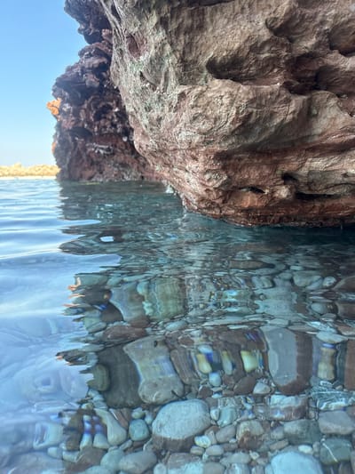 The image depicts a close-up view of crystal-clear water meeting the rocky shore near Sveti Stefan, Montenegro. The water's surface is rippled, creating distorted reflections of the submerged pebbles and the underside of the rock formations. The rocks themselves are rough and textured, with shades of brown and reddish hues. The underwater scene reveals a bed of smooth, rounded pebbles of varying sizes and colors, some appearing lighter due to the refracted sunlight. The sky above is a clear, pale blue. No people or man-made objects are visible in the frame.