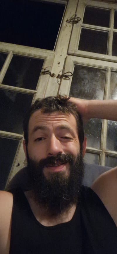 The image is a close-up, selfie-style photograph of a man with dark, unkempt hair, a prominent beard, and a dark t-shirt. He is looking at the camera with a winking expression and his right hand is resting on his head. Behind him is a large, old-fashioned window frame with multiple panes. The window is dirty and the background visible through the panes appears to be dark, suggesting it is nighttime or a dimly lit interior. The overall impression is casual and informal. There is no discernible information in the image to indicate a specific location in the city of Wattenwil, Switzerland.