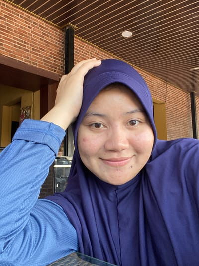 A young woman wearing a dark purple hijab and a blue long-sleeved shirt is taking a selfie. She is indoors, with a brick wall and a wooden paneled ceiling visible in the background. Her left hand is raised and resting on the top of her head. The woman is smiling and looking at the camera. There are some blemishes on her face. The lighting suggests it is daytime.