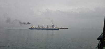 The image captures a wide shot of the sea with a dull, overcast sky. In the foreground, the choppy grey water extends to the horizon. Two large cargo ships are visible in the mid-ground. The ship on the left is a bulk carrier, painted blue and white, with cranes and two smokestacks emitting dark black smoke. The ship on the right is a tanker, painted dark red and black. In the far distance, a long breakwater or pier stretches across the horizon with a small structure, possibly a lighthouse, on its far end. The scene appears to be in the waters near Colón, Panama, given the industrial shipping activity. The overall atmosphere is somber due to the overcast weather and the visible pollution from the ship.