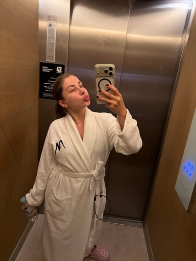An individual is taking a selfie in an elevator. The individual is wearing a white bathrobe and has black nail polish. They are holding a smartphone in their right hand, with which they are capturing the image. The elevator appears to have stainless steel walls and wooden accents. There is a sign on the wall with some text in a foreign language. The elevator buttons are visible on the right side of the image. The floor of the elevator is visible at the bottom. The location is in Žunovnica, Bosnia and Herzegovina.