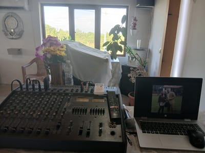 The image shows an indoor setting, likely a room used for audio mixing. Central to the scene is a large, black Montarbo audio mixer with numerous knobs, faders, and input/output jacks. A black Toshiba remote control rests atop the mixer near a wired microphone. Next to the mixer is an open HP laptop, its screen displaying a black and white photograph of a man and a woman. A black mouse is positioned to the right of the laptop.
In the background, a window provides a view of a landscape with trees, possibly indicating a location in the countryside. Part of the scene is obscured by a white tablecloth draped over an unseen object. To the left of the draped object, a flower arrangement in a vase sits on a surface along with a water bottle. A vintage-style clock hangs on the wall in the background on the left side of the window. A water dispenser, along with a potted plant is seen on the right side of the window.
Based on the details and context, the location is possibly a recording studio or an audio production setup in a residence, and given the latitude and longitude, the region is potentially Vetren Dol, Bulgaria.