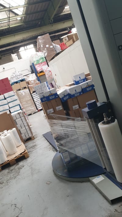 The image shows the interior of a warehouse in Vestec, Czechia. The scene is dominated by stacks of boxes and pallets, some wrapped in plastic. A stretch wrapping machine is visible in the foreground, with a roll of plastic wrap attached. There are no people visible in the image. The warehouse appears to be well-lit, with natural light coming from the ceiling. The floor is concrete, and there are some marks and stains on it. The walls are white, and the ceiling has a metal structure. The overall impression is of an organized, working warehouse environment.