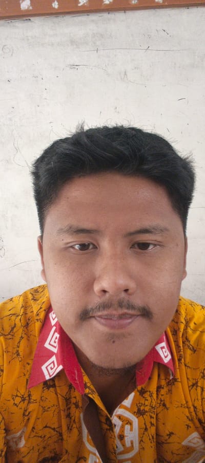 This is a medium shot, close-up selfie of a young man, likely in his early 20s, set against a light-colored, textured wall. The man has dark, messy hair, a prominent mustache, and dark brown eyes. He is wearing a vibrant yellow shirt with a dark, abstract pattern. The shirt has a red collar with a white geometric design, featuring what appears to be a stylized square or maze motif repeated around the neckline. The man's expression is neutral, with his gaze directed towards the camera. The lighting suggests it is daytime, possibly indoors or in a shaded outdoor area, as there are no strong shadows. The textured wall behind him might indicate a simple, unadorned indoor setting, or perhaps an outdoor wall with a rough finish. There is no visible text in the image. The overall impression is a casual, personal photograph. The location context of Sukabumi, Indonesia, places the subject within a specific geographical region, but offers no direct visual cues within the image itself to confirm this.