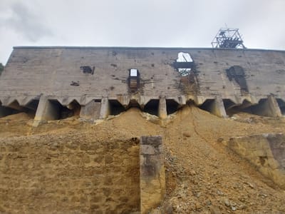 The image depicts a large, derelict concrete structure, likely a former industrial building, situated on a steep, earthy embankment. The concrete wall is weathered and damaged, with several large openings revealing the interior and the sky beyond. The lower section of the structure is supported by a series of concrete arches, beneath which piles of earth and rubble have accumulated. A rudimentary wooden framework, possibly a remnant of a mining headframe, is visible at the top of the structure. The ground is covered in loose soil, rocks, and debris, suggesting an abandoned or disused site. The sky is overcast, casting a diffused light over the scene. The location is identified as the historical mine tailings of Kapan, Armenia. There are no people or animals present in the image.