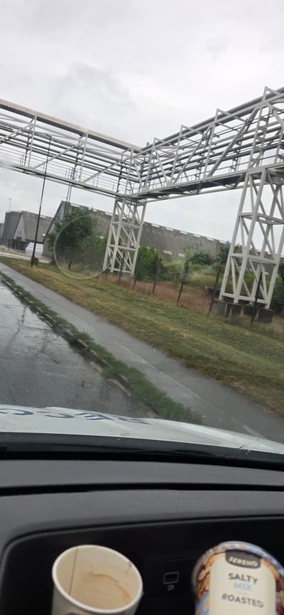 The image captures a view from inside a vehicle looking outwards. The primary subject is an expansive white metal structure, likely part of a larger industrial or infrastructure complex, stretching across the frame. This structure is characterized by its geometric, truss-like design. Beyond the structure, a grassy area and a paved path are visible, suggesting an outdoor setting. The path appears wet, indicating recent rain. A grey, overcast sky dominates the upper portion of the image, hinting at cloudy or rainy weather.
On the right side of the frame, inside the vehicle, a disposable coffee cup with brown residue is placed in a holder. Next to it, a cylindrical container of what appears to be snacks is visible, with text on its label. Partially legible text on the snack container reads "FERRERO," "SALTY MIX," and "ROASTED."
The orientation of the image is rotated, making it appear as if the viewer is looking sideways. The scene likely takes place during the day, given the ambient light. The overall mood is neutral, dominated by the architectural element and the weather conditions. The presence of the coffee cup and snack container implies the ongoing activity of traveling or waiting. The location context of Aarhus, Denmark, adds a geographical reference to the scene.