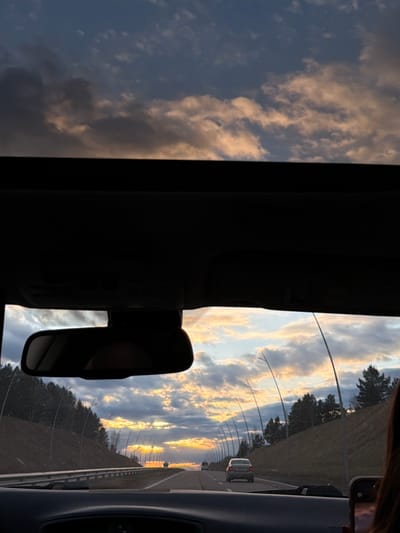 Captured from within a vehicle on a highway, the image shows a view through the front windshield. The sky is filled with clouds illuminated by the setting sun, casting golden hues. The road stretches ahead, lined with trees and lampposts. Other vehicles can be seen in the distance. A rearview mirror is visible within the car. Part of a cell phone and the silhouette of a person's head are visible on the right side. The location is likely near Yemelyanovo, Russia, given the context.