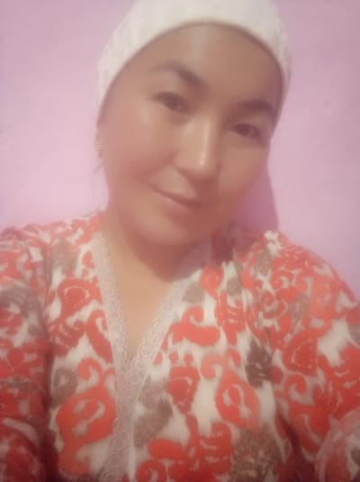 The image is a close-up, selfie-style photograph of a woman wearing a white head covering and a patterned shirt with orange and white floral designs. She is looking slightly to her left, with her mouth partially open. The background is a plain, light pink wall. The lighting is soft, and the image appears to be taken indoors. There are no specific landmarks or contextual clues in the image that would allow for the identification of the city of Kerben, Kyrgyzstan, or any other specific location.