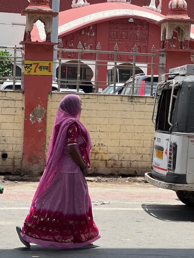 A full-shot of a woman walking on a street in Ajmer, India. The woman is wearing a traditional pink and purple outfit, including a hijab, and is walking from left to right. She is on the right side of the image. Behind her is a brick wall and a red building with intricate carvings. To the right of the woman is the back of a white auto-rickshaw, also known as a tuk-tuk. The auto-rickshaw has a license plate that is partially visible. The street is paved with asphalt and there are some red and white bricks on the side. The sky is visible in the background and it appears to be a sunny day. The area seems to be a busy urban environment with a mix of historical and modern architecture. A sign with Hindi script is visible on a red pillar to the left of the woman.