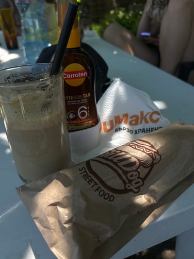 The image shows a collection of items on a white table, possibly outdoors. In the foreground, there is a glass of a frothy, light-colored beverage with a black straw. Next to the glass is a bottle of "Carroten INTENSE TAN TANNING OIL SPRAY SPF 6". To the right of the bottle, there is a white bag with orange text that reads "MAXC" and below it "БЪРЗО ХРАНЕНИЕ". In front of these items, a brown paper bag with a logo depicting a hot dog and the words "STREET FOOD" is visible. The background is out of focus, showing greenery and parts of people. There is no specific indication of the city of Belo pole, Bulgaria in the image itself.