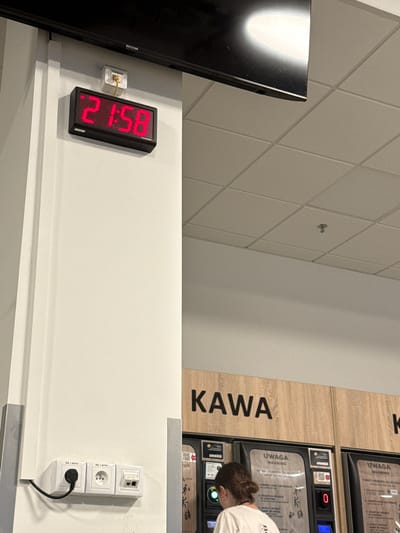 The image depicts a digital clock displaying "21:58" mounted on a white wall. Below the clock, a power outlet with a black plug is visible. To the right of the clock, a vending machine with the word "KAWA" (coffee in Polish) above it is partially visible. A person is standing in front of the vending machine, interacting with it. The environment appears to be indoors, possibly a public space such as a train station or a rest stop, given the presence of the vending machine and digital clock. The lighting suggests an artificial light source. The overall scene is captured from a medium-close shot, focusing on the clock and the vending machine area. Based on the text "Chociule, Poland" provided in the prompt, this scene is located in Chociule, Poland.