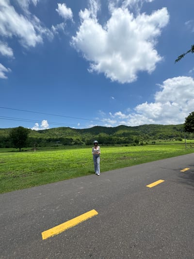A person wearing a white top, blue jeans, and a helmet stands on an asphalt road with yellow lane markings in Ham Thuan Bac, Vietnam. The road curves slightly to the right. To the left of the road is a vibrant green field. In the background, rolling hills covered in lush green foliage rise, and a clear blue sky with scattered white clouds stretches above. Power lines are visible across the horizon.