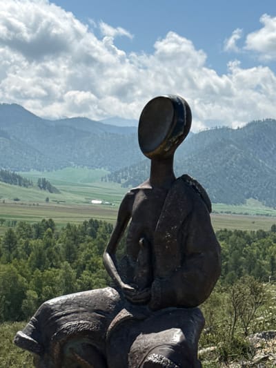 A dark bronze statue of a figure with an abstract, disc-shaped head sits on a rocky outcrop overlooking a valley. The figure is seated, with its torso angled slightly to the left and its head turned to face the same direction. It wears a draped garment over its shoulders and arms. The statue's right arm rests on its lap, holding a small, undefined object. The background features rolling green hills and mountains under a partly cloudy blue sky. A dense forest borders the valley, with fields and a distant structure visible. The statue is situated in the Bichiktu-Boom area of Russia, known for its natural beauty and sculptures.