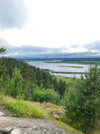 An elevated, wide shot captures a serene natural landscape under a cloudy sky in Khelyulya, Russia. The foreground is dominated by large, moss-covered rocks and a patch of dry grass. A dense evergreen forest stretches across the middle ground, with varying shades of green and the occasional tall pine tree. Beyond the forest lies a tranquil lake, with a thin, man-made structure, possibly a dam or bridge, crossing it. The lake is dotted with small, green islands. The background reveals rolling hills covered in more vegetation and distant buildings, suggesting a rural or semi-rural setting. The overall atmosphere is peaceful and overcast.