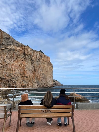 The image displays a coastal scene in Torroella de Montgrí, Spain, featuring three individuals seated on a wooden bench with their backs to the camera. The bench, which has metal supports, shows graffiti on its backrest. From left to right, the individuals are observed wearing a brown, possibly knitted, hat and dark jacket; a dark jacket revealing long, light brown hair; and a black beanie with a multi-colored dark blue and red jacket. They are positioned on a red-brown paved promenade, facing the Mediterranean Sea, which features dark blue water and white breaking waves against rocks and the base of a large cliff. A metal railing separates the paved area from the water. A significant, rugged reddish-brown rock cliff dominates the left side and background, exhibiting visible geological strata and sparse vegetation atop its summit. The sky is partially cloudy with scattered white clouds against a blue background. A fragment of another wooden bench or table is visible to the far left.