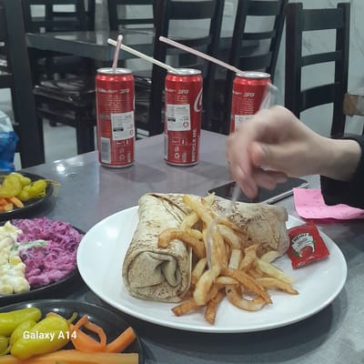 Here is a detailed description of the image:

**Scene:** A dining table setup for a meal.

**Subjects:** A hand is visible near the food, likely belonging to someone dining. There are plates of food and drink cans.

**Objects:**
*   A white plate with a wrap (possibly a shawarma), French fries, and a packet of Heinz ketchup.
*   Three red Coca-Cola cans with straws.
*   Two black plates with salad.
*   A pink piece of paper with some writing on it.
*   The text "Galaxy A14" and "RECYCLE ME"

**Environment:**

*   The image appears to be taken indoors, likely in a restaurant or dining area.
*   The background includes chairs with dark frames.

**Location:** The location can't be determined in the city of Nuseirat, Palestinian Territories without other images.