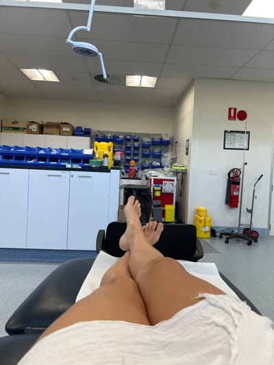 The image presents a first-person perspective from an individual lying supine on an examination table within a medical facility. The specific location in Corrimal, Australia, is not identifiable from the visual content. The subject's bare legs are visible from the upper thighs down, crossed at the ankles, resting on a black examination table covered by a white disposable sheet. A white, crinkled garment partially covers the subject's lower torso.
The background reveals a functional medical environment. Along the left wall, a system of white base cabinets is topped with a blue countertop holding numerous blue plastic storage bins containing medical supplies. Cardboard boxes, including one labeled "Baxter," are stacked above. A yellow sharps container is present on the counter. To the right, a mobile medical trolley with various containers, including another yellow biohazard bin, is visible. On the far right wall, a red fire extinguisher is mounted, positioned next to a wheeled stainless steel IV pole. Another yellow biohazard bin is on the floor to the right of the IV pole. Overhead, a white, multi-LED surgical light is suspended from the ceiling. The ceiling consists of white acoustic tiles with recessed fluorescent light panels and a circular air vent. The floor is a light grey linoleum-style material. No other individuals or active interactions are depicted.