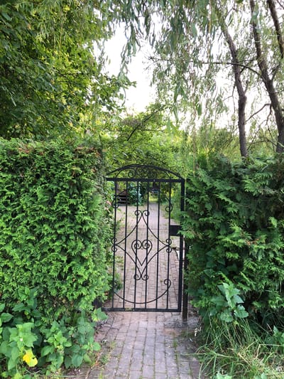 The image depicts a wrought iron gate set within a dense green hedge. The gate features an ornate design with heart shapes and scrollwork. A brick pathway leads through the gate into a garden area beyond. Lush foliage, including trees with drooping branches and other leafy plants, surrounds the gate and pathway. The scene suggests a private garden or backyard entrance. The location is Makeikha, Russia.