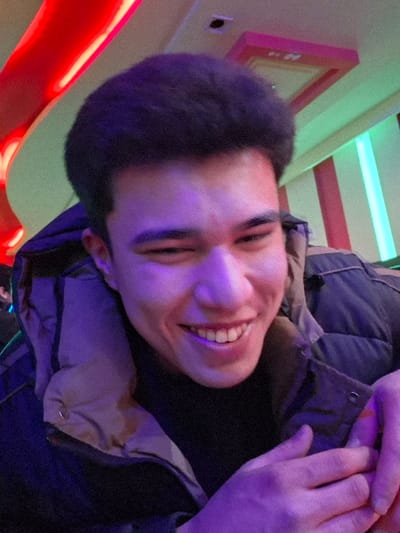 A young adult male, appearing to be in his late teens or early twenties, is depicted in a close-up, chest-up portrait. He has short, dark, curly hair. The subject is smiling broadly, with his left eye (viewer's right) closed or significantly squinted, and his right eye (viewer's left) partially open, exposing his teeth. He is wearing a dark, possibly navy or black, puffer jacket with a light brown or tan-colored inner collar, over a visible black undergarment. His hands are clasped together at the bottom right of the frame, with his left hand gripping his right wrist. A faint orange glow is observable between the fingers of his right hand. The background is an indoor environment characterized by vivid artificial lighting, including a prominent curved red light fixture at the top left, a rectangular pink ceiling fixture at the top right, and vertical green and pink/red strip lights on a wall. The subject is predominantly illuminated by a purple light source. The precise location within the city of Takhtakuprik, Uzbekistan, cannot be determined from the visual information provided; however, the lighting and fixtures suggest an entertainment venue such as a karaoke bar, club, or brightly lit restaurant.