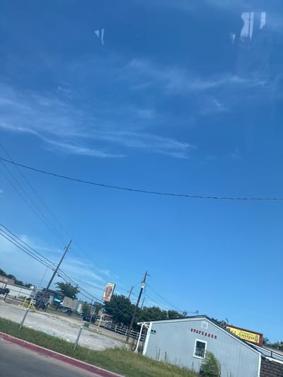 The image shows a daytime exterior scene in Balch Springs, Texas, USA. The sky is a clear blue with scattered wispy clouds. In the foreground, there is a patch of green grass and a red-lined curb. Power lines crisscross the scene, supported by utility poles.

A light blue building with a peaked roof is visible, bearing the word "PROFESSOR" in red letters above a window. Adjacent to this building, a yellow sign with red lettering reads "Taqueria El Amigo MEXICAN FOOD." Several vehicles are parked in a lot in front of the buildings. In the background, there are trees and other structures, including what appears to be an AT&T store. The overall impression is of a commercial area on a sunny day.