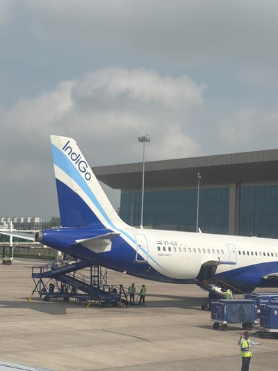 An Indigo Airlines airplane, registered as VT-ILZ and an Airbus A321neo, is parked on the tarmac at an airport. The aircraft's tail, adorned with the Indigo logo, is visible against a cloudy sky. A jet bridge or airstair is connected to the airplane, and several ground crew members are present. Some individuals are near the aircraft and the airstair, and one person in a high-visibility vest is directing traffic with illuminated batons. Two blue baggage carts are positioned near the aircraft. In the background, a modern airport terminal building with large, reflective windows and a gray overhang is visible. The scene is likely at an airport near Vikram Nagar, India, given the presence of Indigo Airlines, an Indian carrier.