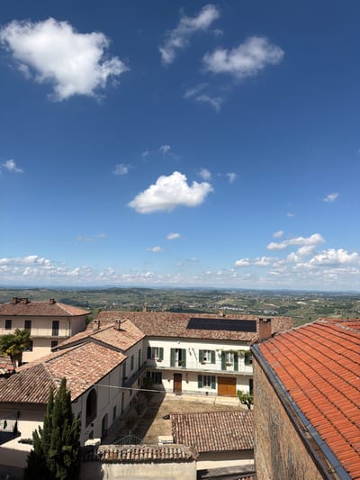 View of Castiglione Tinella, Italy, from above. The image shows a clear blue sky with white cumulus clouds. Below, the rooftops of buildings with red-orange terracotta tiles are visible. In the background, a vast expanse of green and brown hills and valleys are partially obscured by clouds. A white building with green shutters and a brown door is centrally located. Solar panels are installed on the roof of an adjacent building. The overall impression is of a rural landscape in sunny weather.