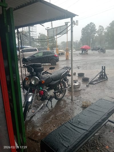 The image captures an outdoor scene under heavy rainfall in Kapkures ward, Kenya, on 2026/02/28 at 17:29. The environment is saturated, with large puddles and muddy areas covering the ground.
In the foreground, under a corrugated metal roof, a black motorcycle featuring a circular headlight, chrome front fender, and orange turn signals is parked on its kickstand. Two black tires are stacked vertically behind the motorcycle. To the right, a black, rectangular padded bench or cot lies wet on the ground. A green and red vertical structure, possibly a door frame, borders the left edge of the frame. Water visibly drips from the edge of the corrugated roof.
Beyond the shelter, the ground remains wet and uneven. A dark-colored hatchback car is parked to the left of a vertical metal pole that supports overhead cables. Further into the scene, three individuals are clustered together under a large red umbrella, engaged in an unidentified activity on the wet ground. To their right, a second motorcycle is parked. Scattered on the ground are several silver-colored vehicle wheel rims and a black metal frame structure. The mid-background features multiple utility poles and large, blank billboards.
The background shows a line of trees beneath an overcast, grey sky, indicative of ongoing heavy precipitation. Additional vehicles are partially visible in the distance.