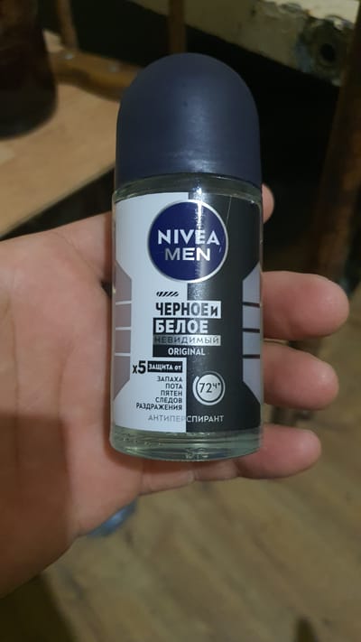 A person is holding a NIVEA MEN roll-on deodorant. The deodorant is black and white with Russian text. The text says "ЧЕРНОЕ БЕЛОЕ" (Black White) and "НЕВИДИМЫЙ ORIGINAL" (Invisible Original). It also mentions "ЗАЩИТА ОТ ЗАПАХА, ПОТА, ПЯТЕН, СЛЕДОВ РАЗДРАЖЕНИЯ" (Protection from odor, sweat, stains, irritation). There is also a "72h" symbol indicating 72-hour protection. The background is blurry and appears to be an indoor environment, possibly in Karankul, Uzbekistan.