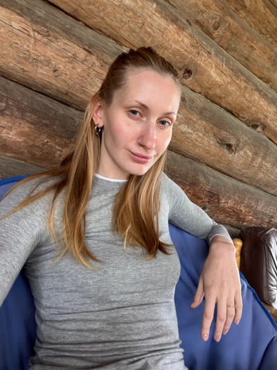 A young Caucasian woman with long, reddish-blonde hair pulled back is seated and looking towards the camera. She is wearing a grey long-sleeved shirt. She has a nose ring and hoop earrings. Her left arm is resting on her lap, with her hand visible. She is seated on a blue surface, possibly a cushion or chair, against a backdrop of weathered wooden logs. The environment suggests an outdoor setting, potentially a rustic cabin or porch, within the city of Merkuriy, Russia. The lighting appears natural and diffused.